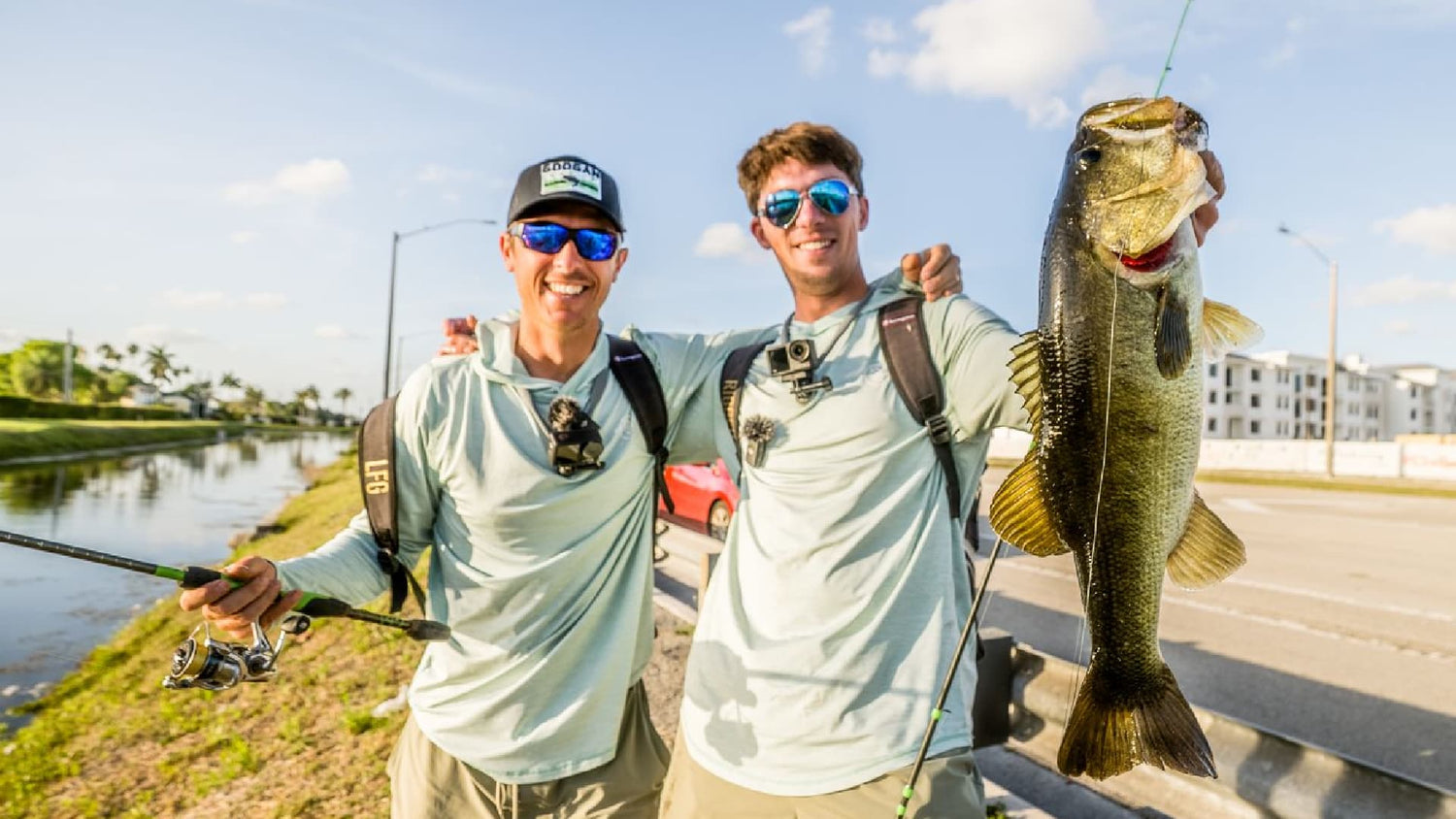 Two anglers bank fishing, holding a largemouth bass beside canal