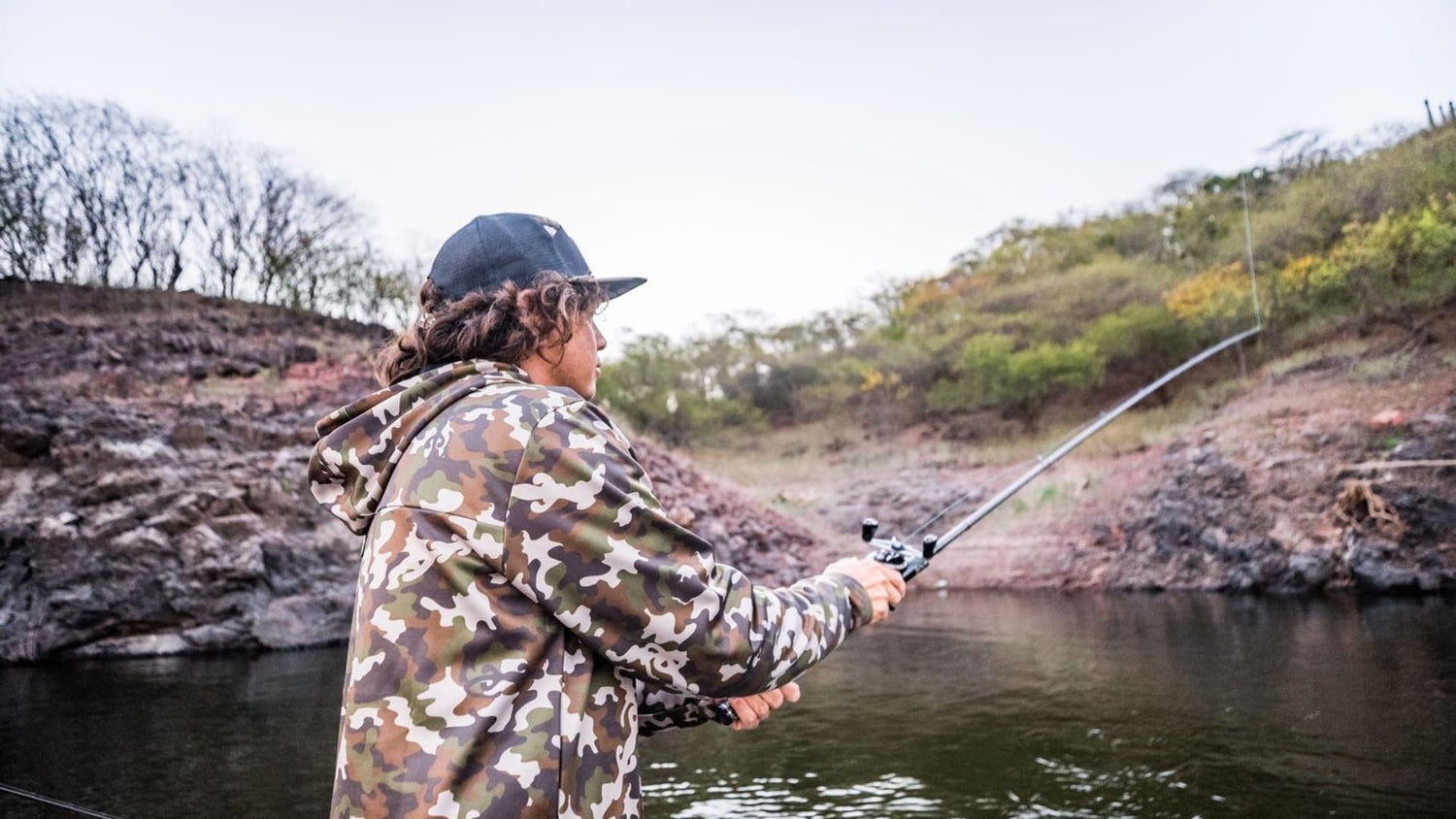 Angler in camo pitching a baitcaster towards rocky shoreline cover.